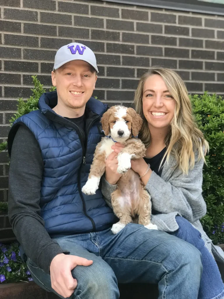 Two people sitting together, holding a small dog with light brown and white fur; background includes a brick wall and green bushes.