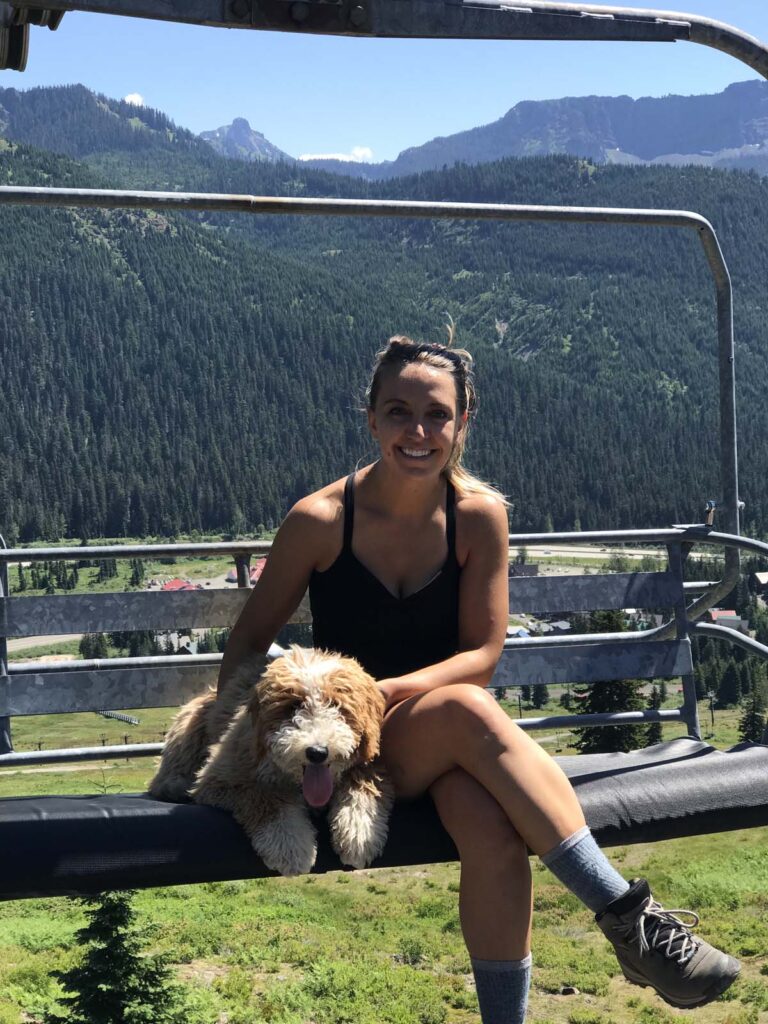 Dr. Chloe Mirick sitting on a ski lift with a fluffy light brown and white dog,Jerry. The background features a scenic view of mountains, green forests, and clear blue skies.