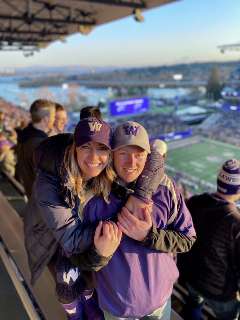 Fans in a stadium wearing purple 'W' caps, watching a sporting event during the day.