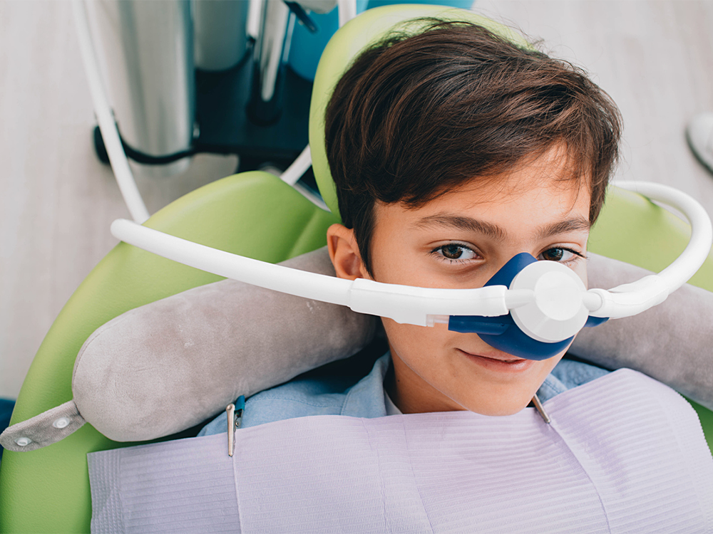 The image shows a person sitting in a dental chair, wearing a dental bib, with a gray neck pillow behind their head. A white dental instrument, likely a bitewing X-ray holder, is positioned around their head. The chair is green, and there are dental tools and equipment visible in the background.