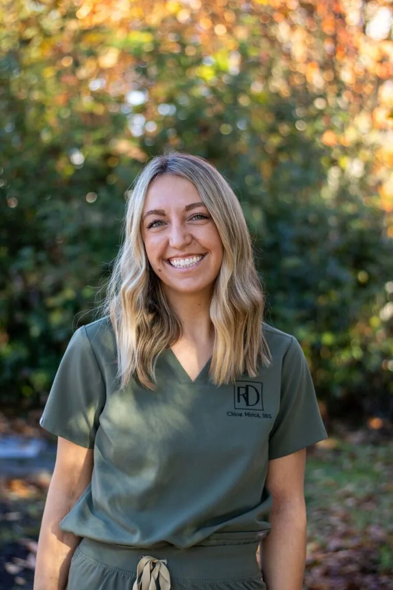 A woman wearing a green scrub top embroidered with the name Chloe Mirick, DDS' stands outdoors in front of greenery and autumn-colored leaves.