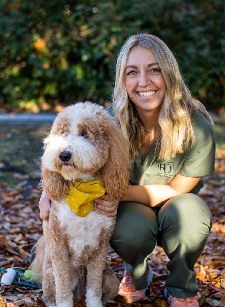 Dr. Chloe Mirick in green scrubs with long blonde hair kneels next to a fluffy, curly-haired Jerry wearing a yellow bandana, set against a background of greenery and fallen leaves.