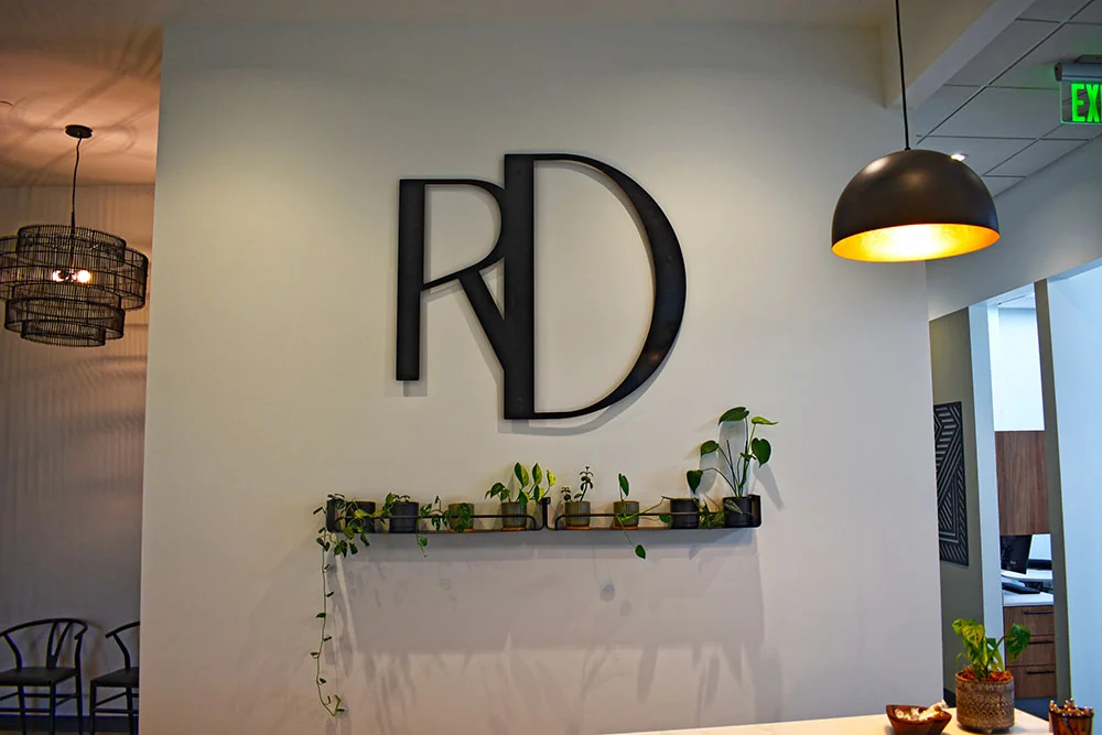 Interior with a large black "RD" logo on the wall above a shelf holding small potted plants; modern lighting fixtures on either side.