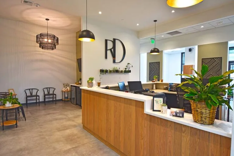 Interior design with a large black "RD" logo on the wall above a shelf holding small potted plants; modern lighting fixtures on either side and countertops that could be a reception area.