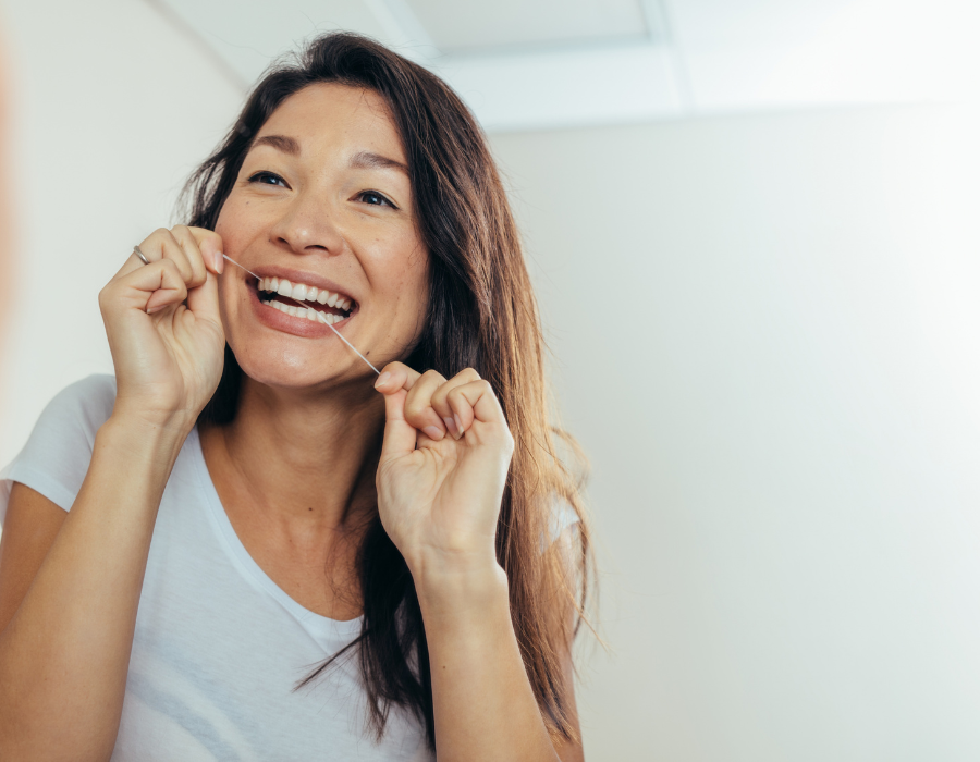 The image shows a person with long hair wearing a white shirt. She was flossing her teeth with both hands. The background is an indoor setting with a light-colored wall and ceiling.