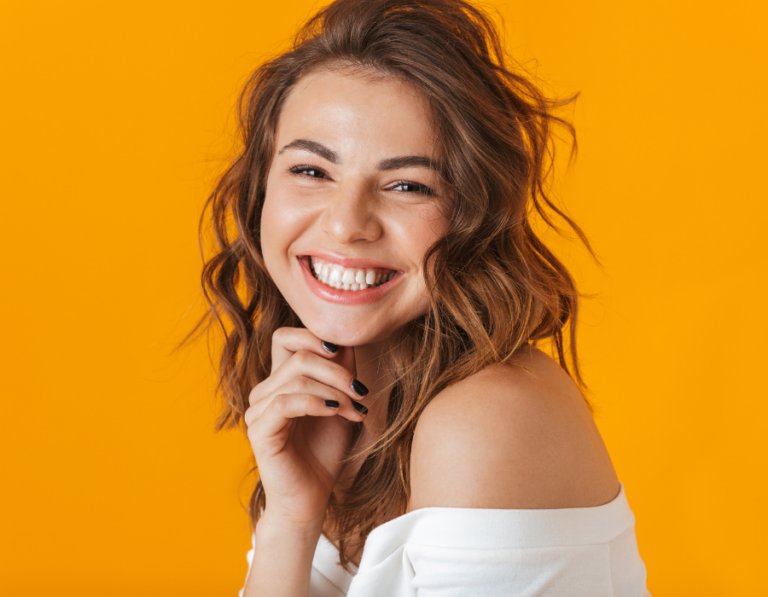 A woman smiling and holding her chin, wearing an off-shoulder white shirt in an orange background.