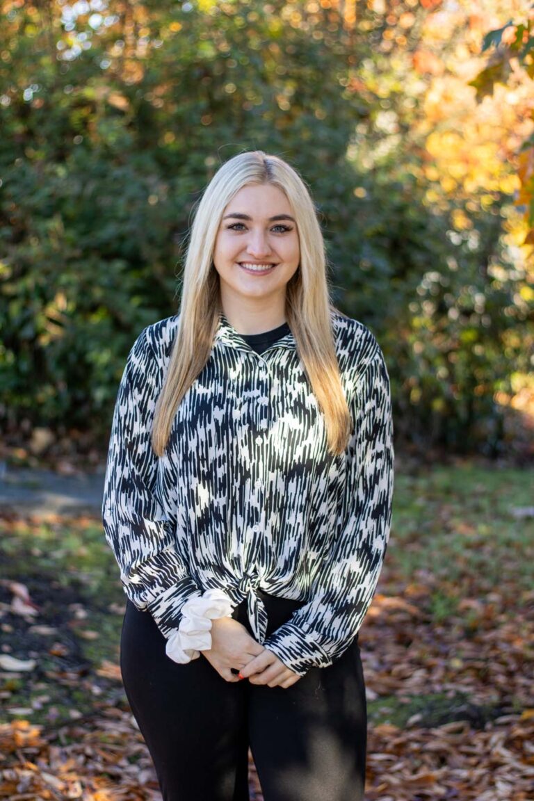 A woman standing outdoors in a natural setting with trees and foliage in the background, wearing a black and white patterned long-sleeve blouse tied at the waist and black pants. The ground is covered with fallen leaves, indicating it is likely autumn.