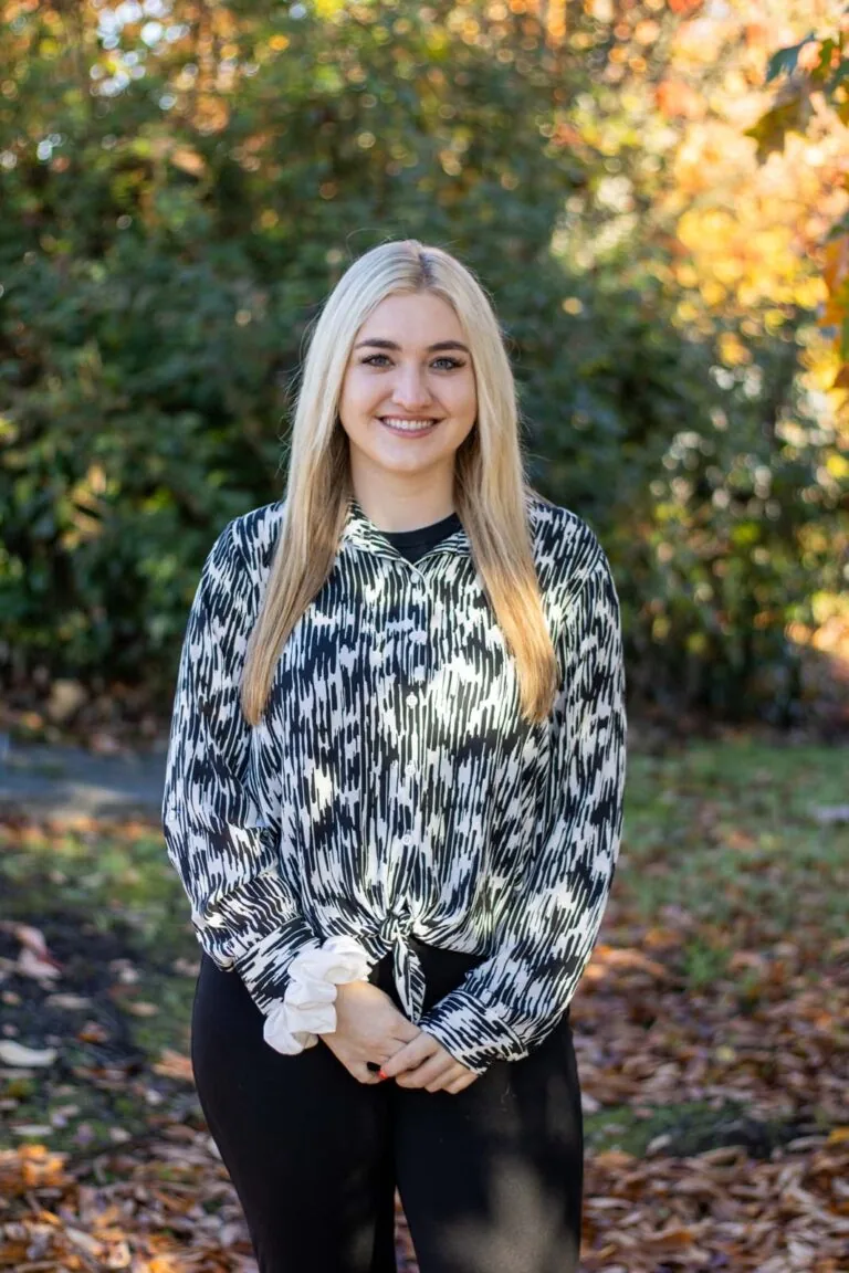 A woman standing outdoors in a natural setting with trees and foliage in the background, wearing a black and white patterned long-sleeve blouse tied at the waist and black pants. The ground is covered with fallen leaves, indicating it is likely autumn.