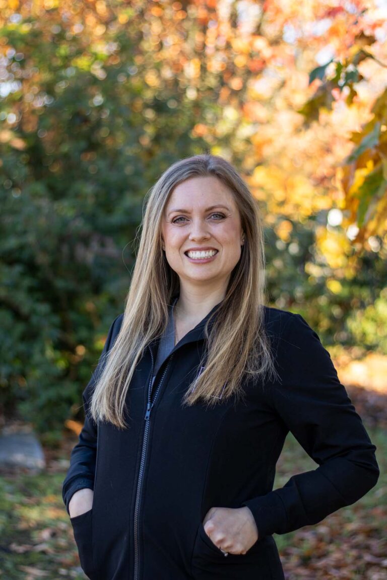 A woman standing outdoors, wearing a black jacket. The background is green foliage with autumn-colored leaves, indicating a fall season setting.