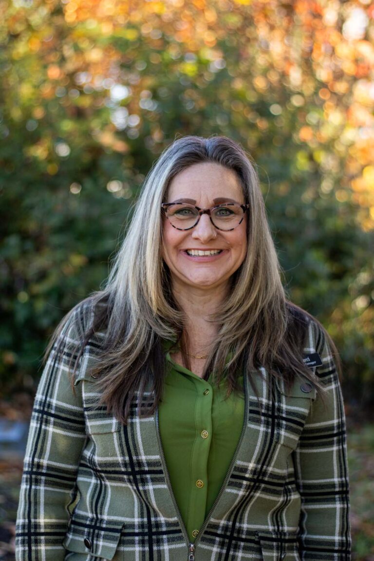 A woman standing outdoors wearing a green shirt and a plaid jacket. The background consists of greenery with autumn-colored leaves.