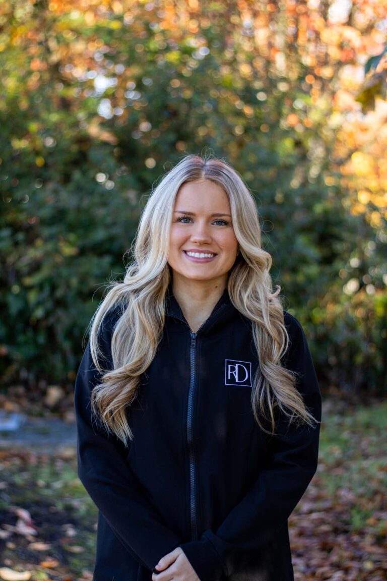 A woman standing outdoors, wearing a black jacket with an 'RD' logo on the left side of the chest. The background is green foliage with autumn-colored leaves, indicating a fall season setting.