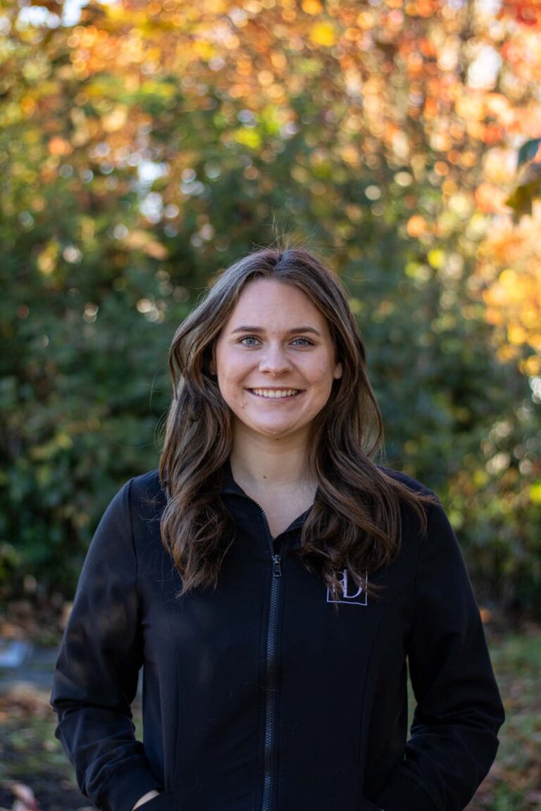 A woman standing outdoors, wearing a black jacket with an 'RD' logo on the left side of the chest. The background is green foliage with autumn-colored leaves, indicating a fall season setting.