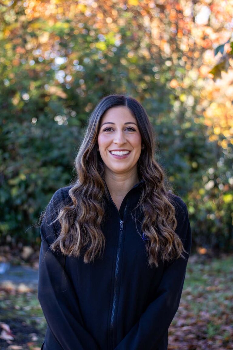 A woman standing outdoors, wearing a black jacket. The background is green foliage with autumn-colored leaves, indicating a fall season setting.