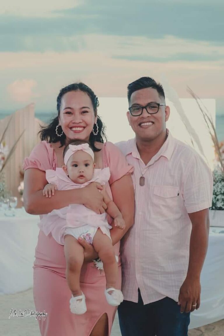 Three people standing outdoors, one holding a baby; background includes a table with floral decorations and a partly cloudy sky.